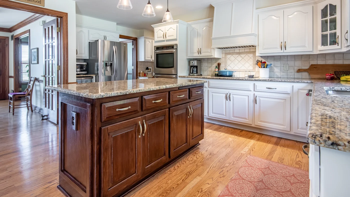 A recently remodeled residential kitchen with light wooden panel floors, a darker-stained island, stone countertops, white tiles backsplash, and kitchen cabinets painted a London Clay color.