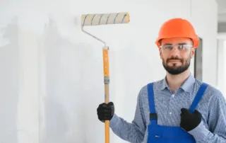 A commercial painter holding a paint roller and wearing a hard hat and gloves in a building where the walls are being painted white.