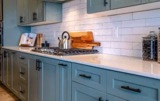 The wall section of a residential kitchen shows new countertops and recently painted cabinets with dark hardware, a white tile backsplash, two filled cookie jars, and the stove top with a tea kettle.
