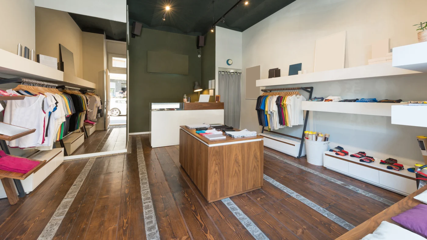 A small retail clothing store with old wooden panel flooring, lots of shelving and hangars on both sides, a new paint job, basic colorful clothing, a fitting room, and a large mirror.