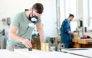 Two men working in a prefinishing and finishing workshop, one applying product to a piece of sanded wood.