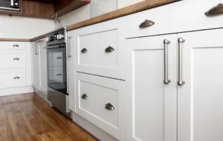 Close-up of newly-painted rich white kitchen cabinets with new hardware, the built-in oven, shiny wood panel flooring, and wood countertops.