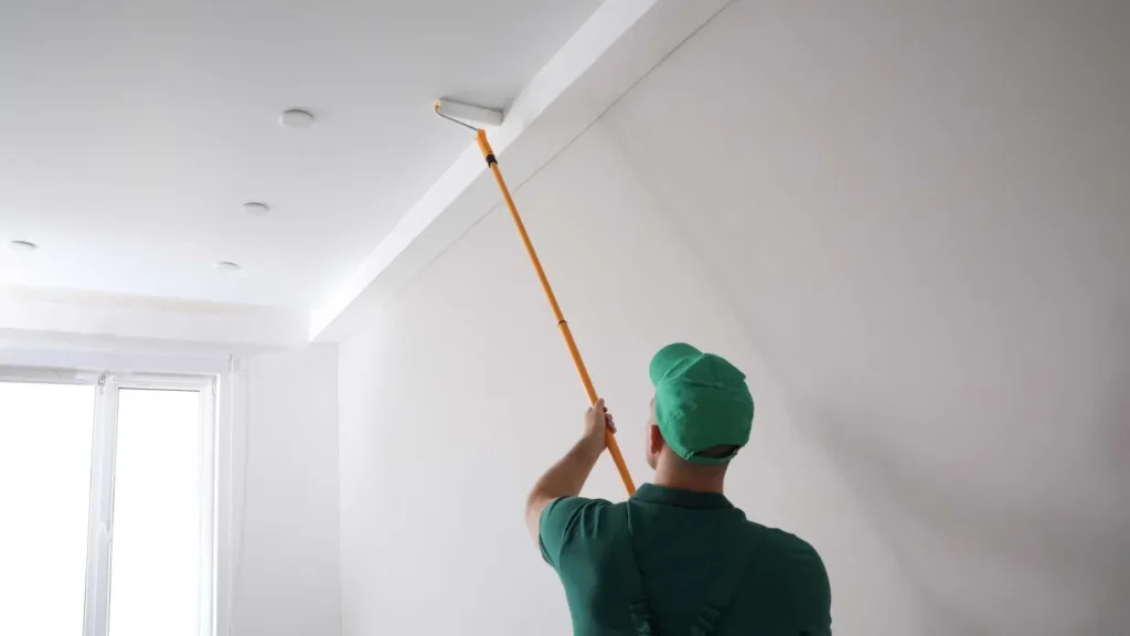 Colorwheel Painting Washington County Painter At Work A professional wall and ceiling painter at work applying a coat of white paint to a ceiling wearing a hat and polo shirt.