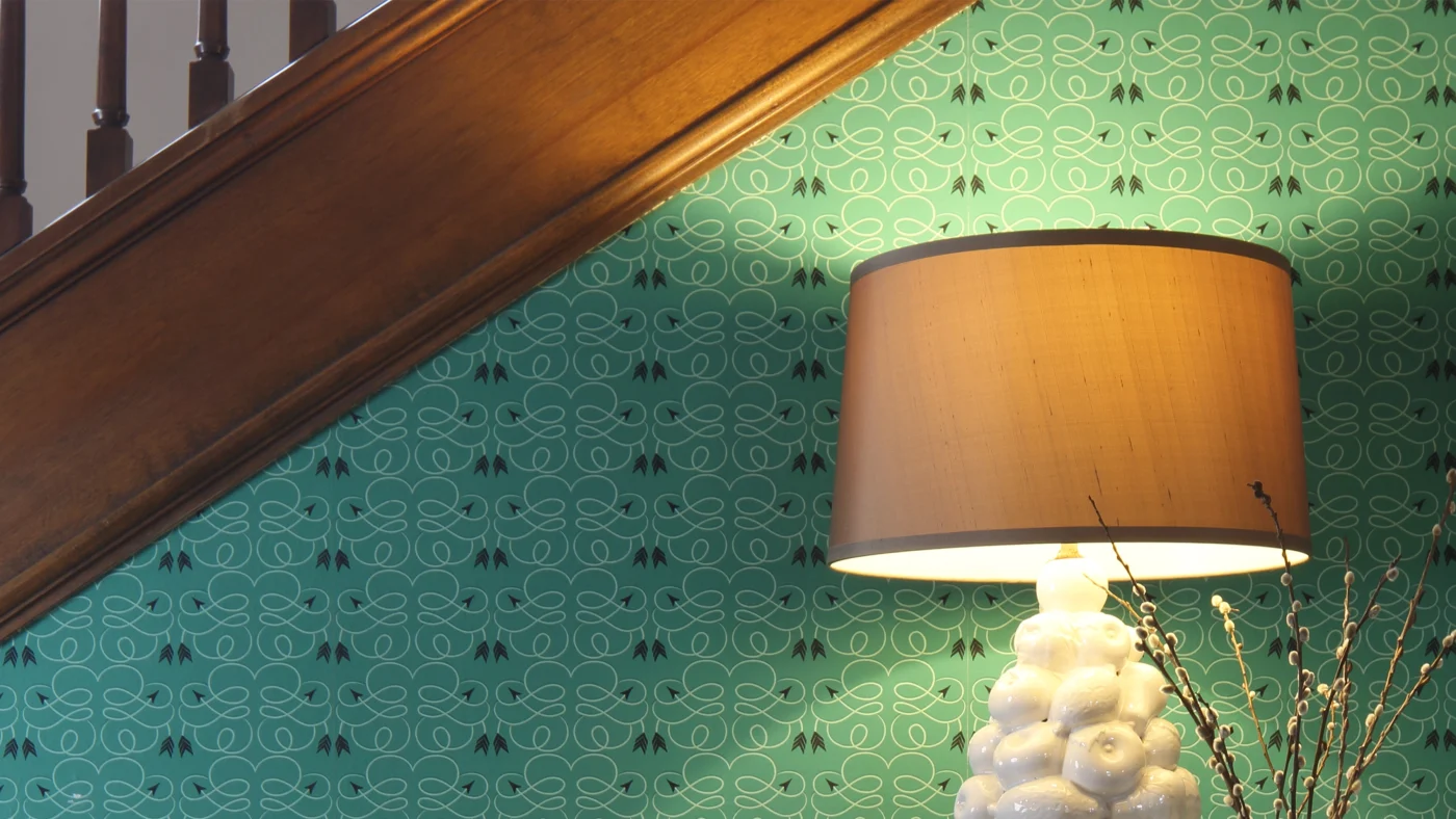 A small accent wall underneath a stairway with a subtle wallpaper design filling the space, and a small classy white lamp sitting in front.