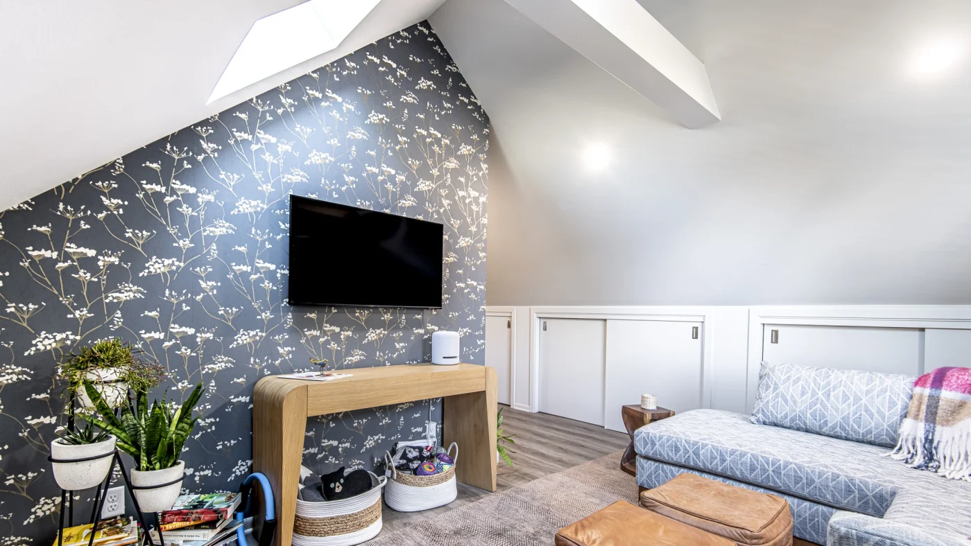 A remodeled attic living space with a wall-mounted TV in front of new floral wallpaper, green flowers in white pots, a comfortable white couch, books and magazines and a skylight.