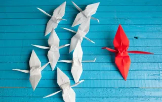 7 white origami birds face a single red origami bird that stands out as a leader among the pack